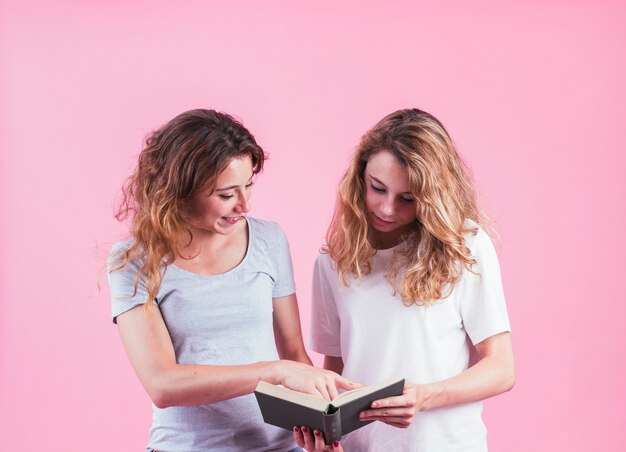 Two female friends reading book against pink background