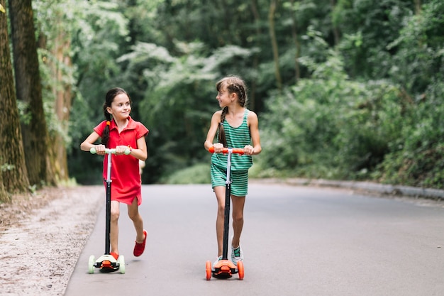 Two female friends riding push scooters on street