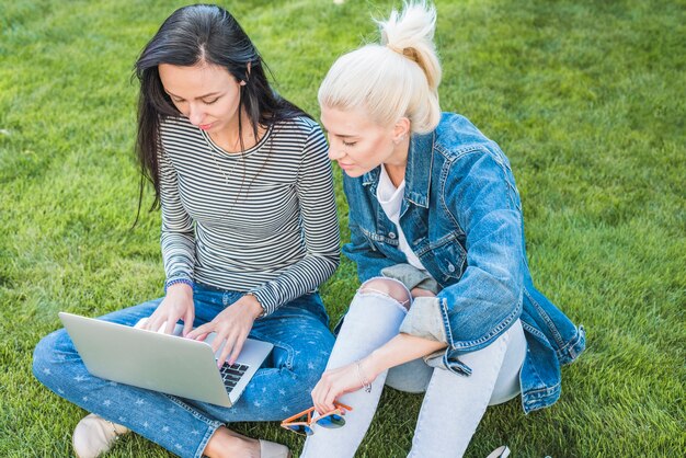 Two female friends sitting on green grass using laptop