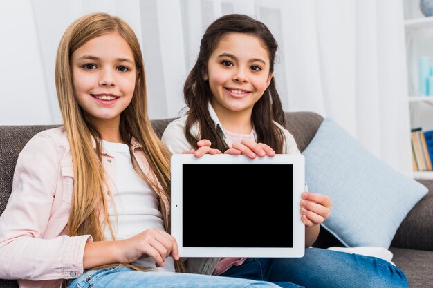 Two female friends sitting on sofa showing blank screen digital tablet toward camera