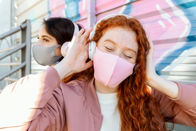 Two female friends with face masks outdoors listening to music on headphones