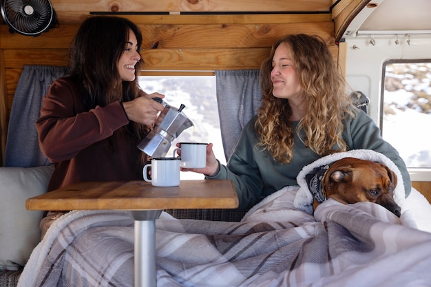 Due amanti femminili che bevono caffè e trascorrono del tempo con il loro boxer in un camper durante il viaggio invernale