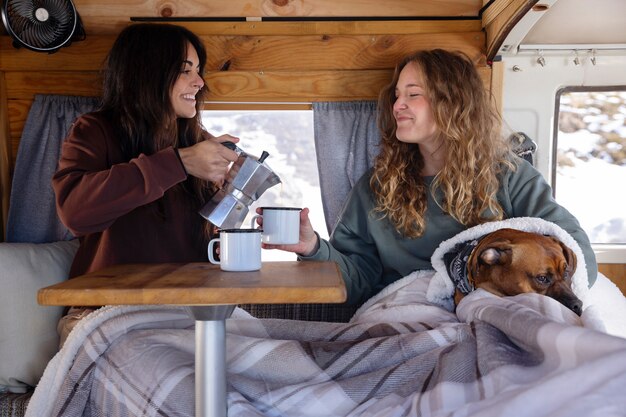 Two female lovers drinking coffee and spending time with their boxer in a camper van during winter trip