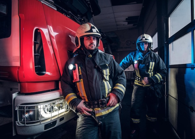 Двое пожарных в защитной форме стоят рядом с пожарной машиной в гараже пожарной части.