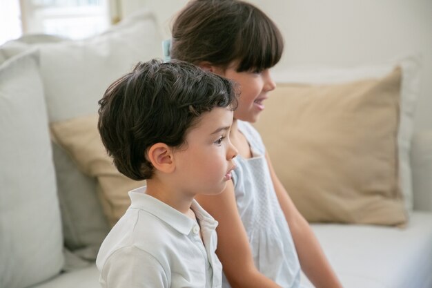 Двое сосредоточенных детей смотрят телевизор дома, сидят на диване в гостиной и смотрят в сторону.