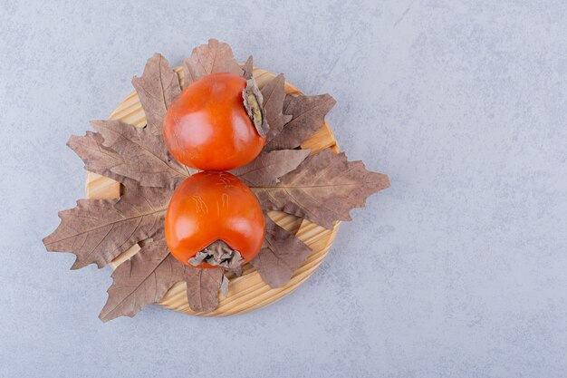 Два свежих плода хурмы и сушеные листья на деревянной тарелке.