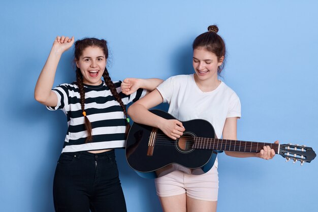 Two friends singing and dancing isolated, lady with playing guitar, winsome girl in striped t-shirt and pigtails raising hands up.