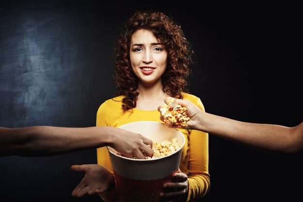 Two friends taking popcorn from girl's bucket. Negative facial emotions