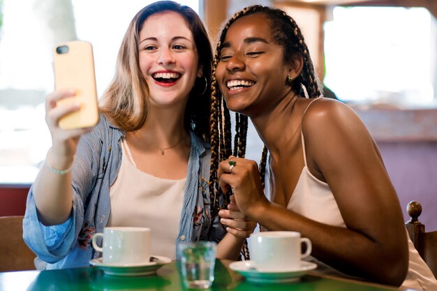 Two friends taking a selfie with mobile phone while drinking a cup of coffee in a coffee shop. Friends concept.