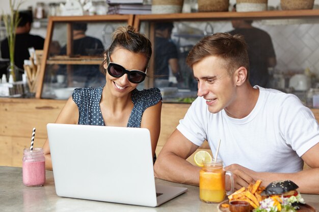 Two friends using laptop pc together during meeting at coffee shop, sitting at table with food and drinks in front of generic notebook computer, looking at screen and smiling