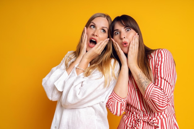 Two funny best friens woman, shoked crazy surprised emotions, studio yellow background.