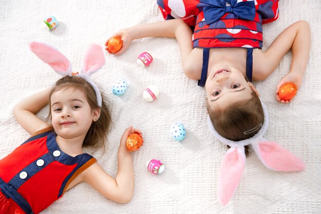 Две забавные милые девочки с пасхальными яйцами и заячьими ушками в красивом ярком платье.