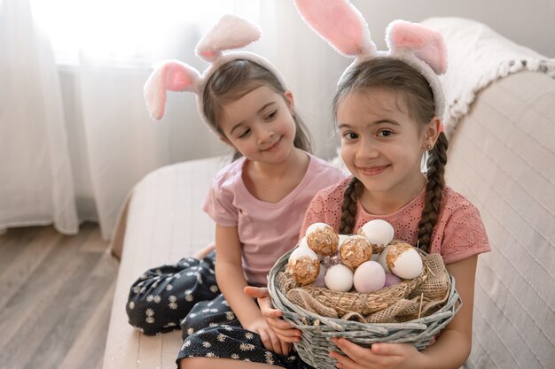 Due bambine divertenti in orecchie da coniglio a casa sul divano con un cesto di uova di pasqua