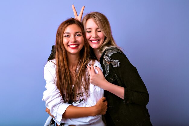 Two funny pretty sisters having playful mood and having fun together, showing finger to camera, casual stylish clothes, purple background.