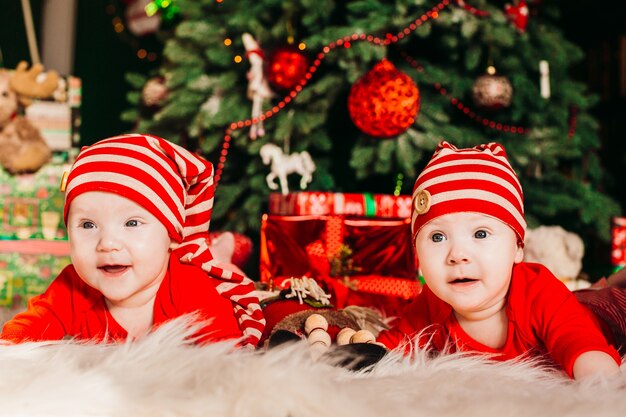 Два веселых близнеца в красных костюмах лежат перед богатой рождественской елкой