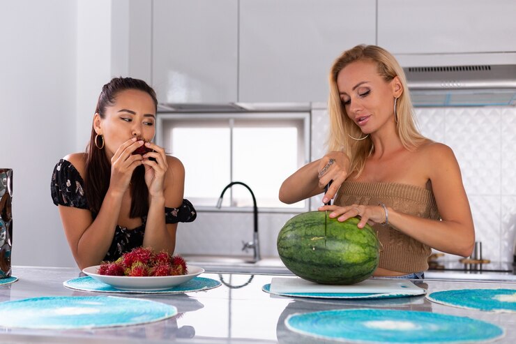 Free Photo two girl friends eating watermelon and rambutan tropical fruits in kitchen
