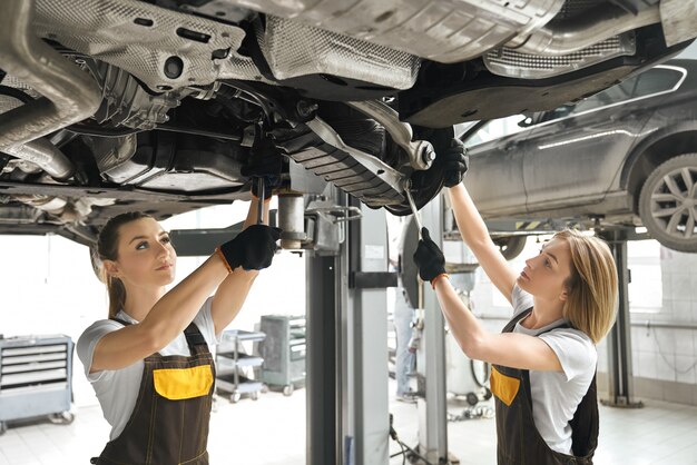 Two girls fixing lifted auto undercarriage, using wrenches.