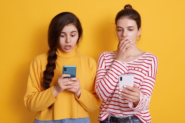 Two girls friends hold phones in hands and looking at screens with big eyes