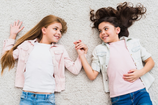 Two girls lying on white carpet holding hand looking at each other