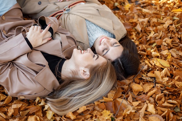 Two girls lying on the yellow leaves at the autumn park. Portrait of female friends. Two girls wearing beige coats.