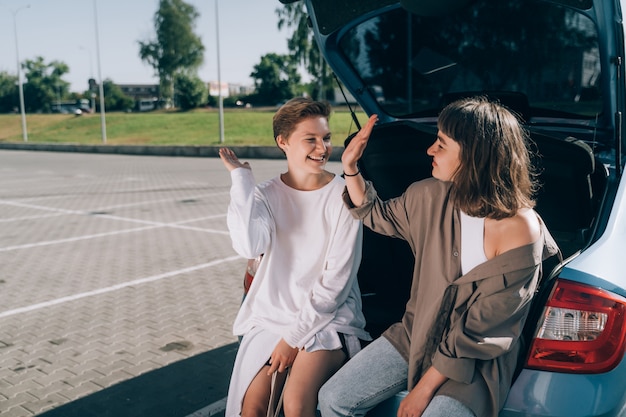 Two girls in the parking lot at the open trunk posing for the camera.