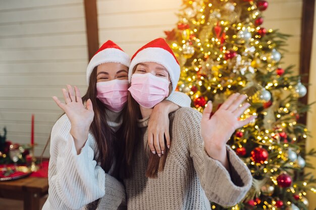 Two girls in protective masks looking at the camera and waving. Christmas during coronavirus, concept