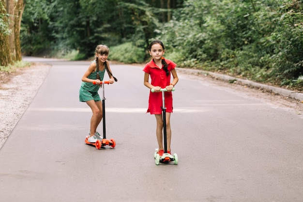 Two girls riding scooters on straight street