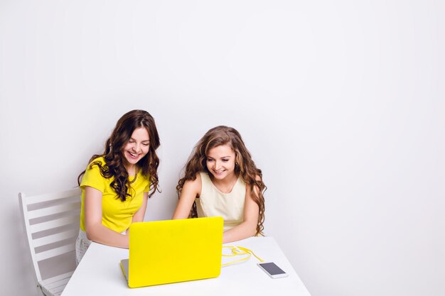 Two girls sit behind a laptop in yellow case. There is a smartphone on the table charging via yellow cable. Both girls are smiling and looking at the screen. One wears yellow t-shirt, another - dress.
