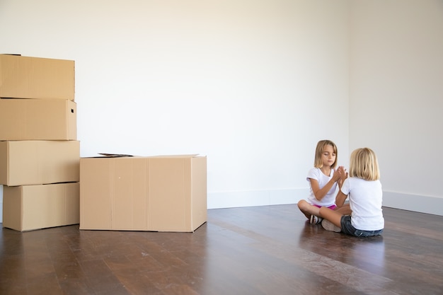 Two girls sitting on floor near heap of boxes in their new apartment and playing together