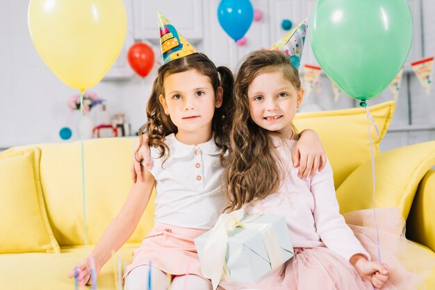 Two girls sitting on yellow sofa holding yellow and green balloons in hands