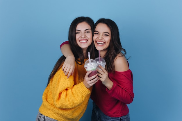 Two girls stand side by side and smile while they drink milkshake and a cocktail