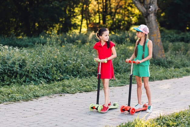 Two girls standing on push scooter looking at each other in the park