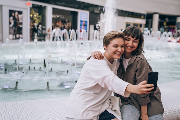 Две девушки делают селфи в торговом центре, фонтан на заднем плане