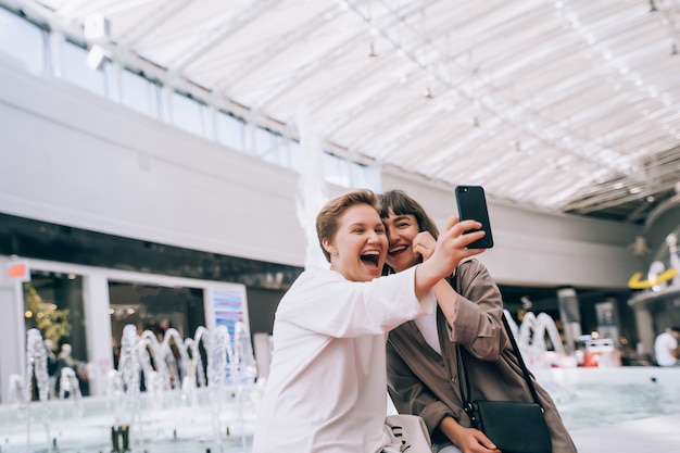 Two girls take a selfie in the mall, next to a fountain