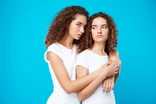 Two girls twins embracing over blue wall