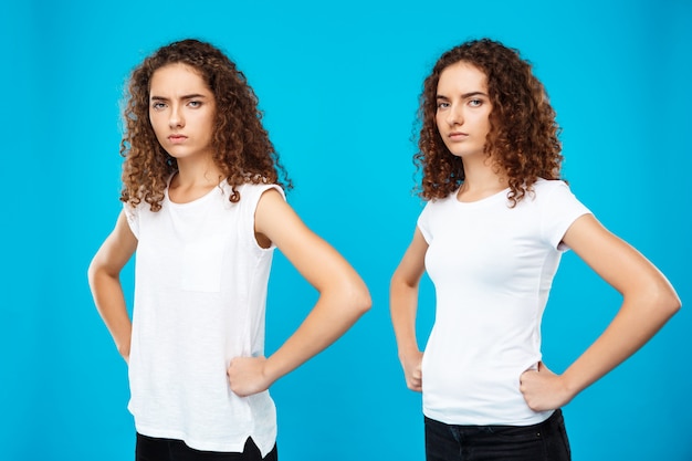 Free photo two girls twins posing with arms akimbo over blue wall