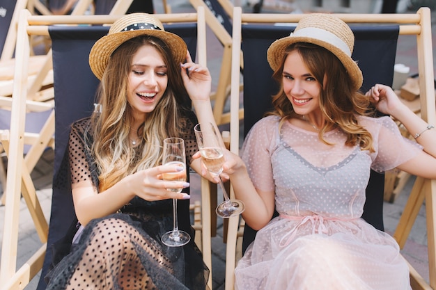 Due ragazze felici con gli stessi cappelli di paglia che celebrano le vacanze con champagne che si rilassano sulle sdraio