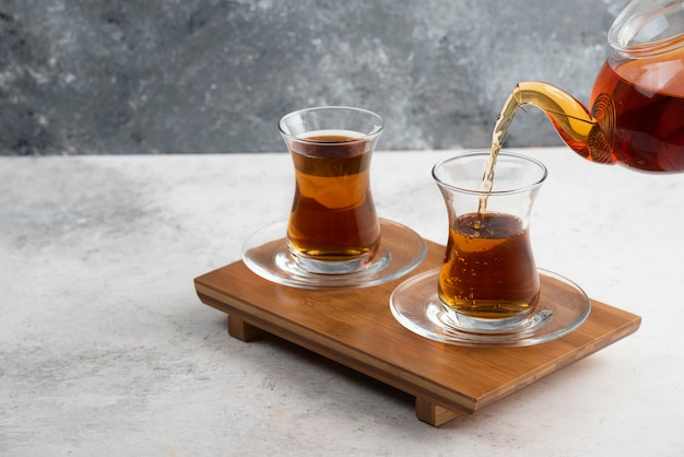 Two glass cups of tea with teapot on wooden board.