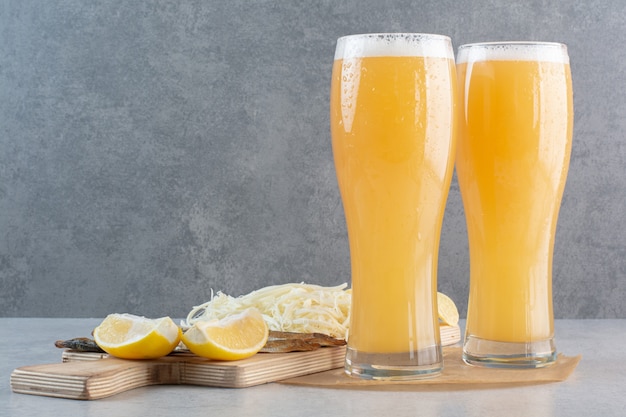 Two glasses of beer with cheese and sliced lemon on gray.