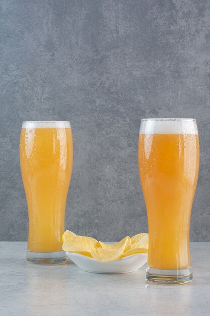 Two glasses of beer with potato chips on gray.