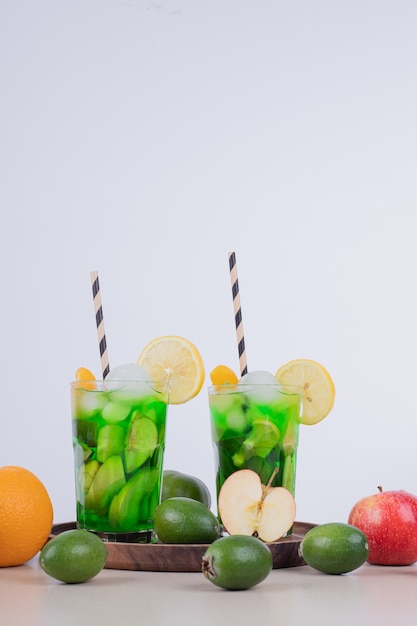 Два стакана сока с кусочками фруктов и свежими фруктами на белой стене.