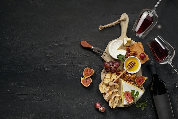 Two glasses of red wine and a tasty cheese plate with fruit, grape, nuts and toasted bread on a wooden kitchen plate on the black stone background