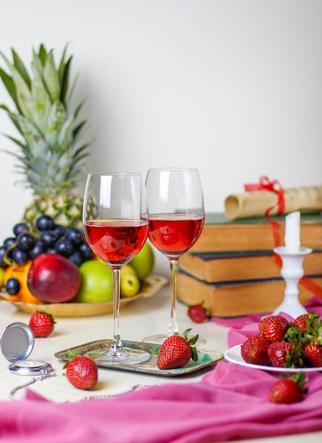 Two glasses of rose wine on white wooden table with vintage books and clock,different tropical fruits and strawberries