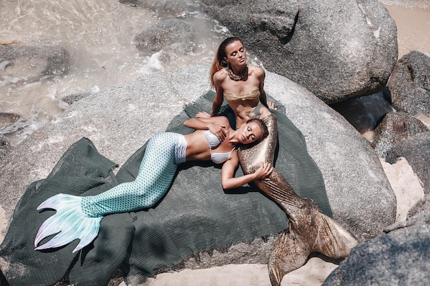 긴 꼬리를 가진 두 명의 멋진 인어가 바위 위에 누워 해변에서 포즈를 취하고 뜨거운 태양 아래 일광욕을 하고 있습니다