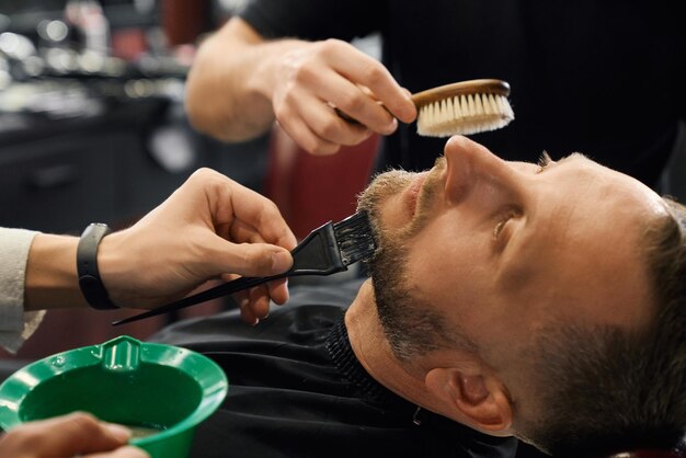 Два парикмахера расчесывают и окрашивают бороду клиента