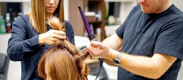 Два парикмахера используют щипцы для завивки длинных каштановых волос клиентов в салоне красоты