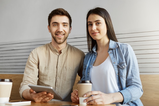 two handsome freelancers sitting in coworking space, drinking coffee and talking about team project. man and woman smiling, , posing for article.