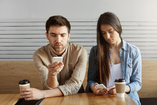 Two handsome students tired after study, sitting in cafeteria , drinking coffee in silence, looking through social networks on smartphones.