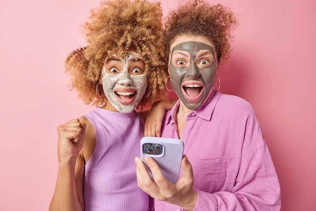 Free photo two happy amazed european women get positive news clench fist holds mobile phone apply beauty masks stare impressed isolated over pink background facial care and cosmetic procedures concept