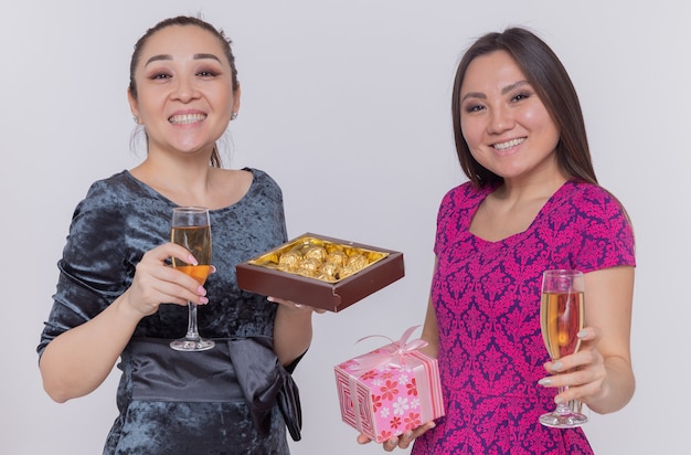 Two happy asian women holding glasses of champagne chocolate candies and present celebrating international women's day smiling cheerfully standing over blue wall